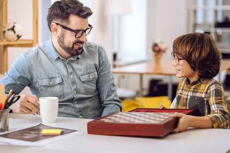 Time for chess. Little handsome boy holding big box with chess looking at his father that is drinking teaの写真素材