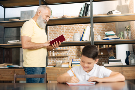 Smiling schoolboy doing homework while his grandfather readingの写真素材