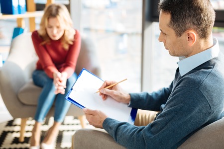 Professional therapy. Sad handsome nice man holding his notes and writing in tem while listening to his patientの写真素材