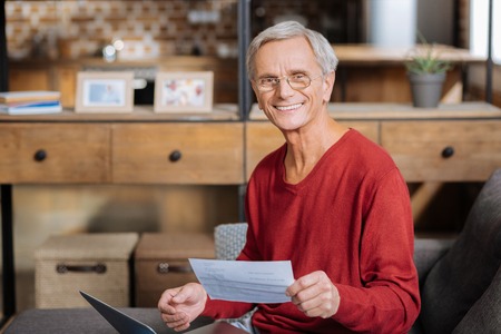 Positive smile. Cheerful delighted positive man holding a piece of paper and looking at you while smilingの写真素材