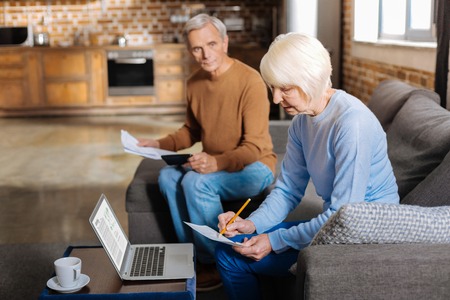 Money and finance. Cheerful nice positive woman smiling and taking notes while planning the family budgetの写真素材