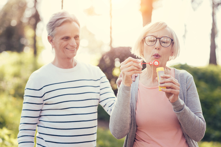 Pleasant activity. Delighted smart carefree woman holding a soap bubbles bottle and blowing bubbles while enjoying the activityの写真素材