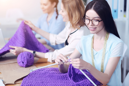 Pleasant work. Cheerful talented enthusiastic women sitting at the table and knitting big purple pieces of clothesの写真素材