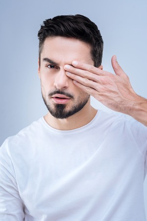 Poor eyesight. Calm attentive serious young man looking not glad while closing his right eye and realizing the bad condition of his eyesightの写真素材