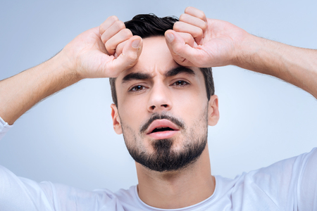 Expressive. Young handsome worried man standing against the blue background and looking emotional while pressing his fists to the foreheadの写真素材