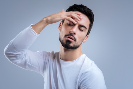 Feeling unwell. Handsome young tired man feeling unwell and having a headache while standing with his eyes closed and touching his foreheadの写真素材