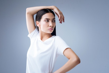 Calm ballerina. Serious attentive beautiful ballerina standing against the blue background and looking at her fingers while her arm being on her headの写真素材