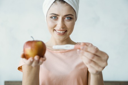 Eat vitamins. Positive delighted woman holding one apple in right hand and demonstrating pregnancy test while looking forwardの写真素材