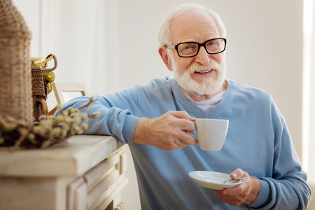 I am relaxed. Delighted pensioner holding cup and keeping smile on his face while looking at cameraの写真素材