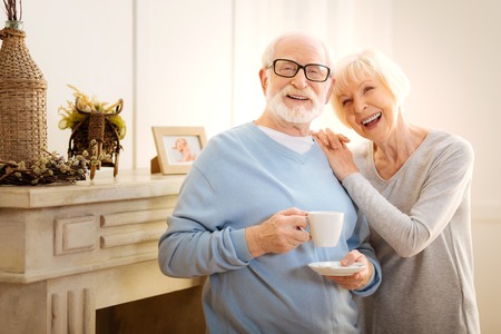 Funny joke. Attractive pensioner keeping smile on his face and holding cup while looking forwardの写真素材