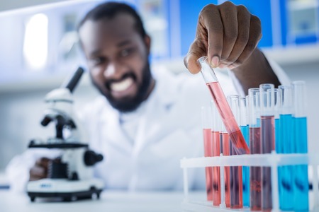 Modern laboratory. Happy smart nice scientists sitting in front of the microscope and taking a test tube while doing an analysisの写真素材