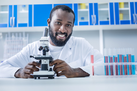 Favourite job. Happy handsome professional scientist sitting at the table and looking at you while being happy about his jobの写真素材