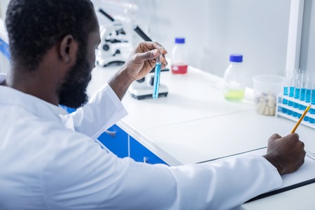 New cure. Nice smart handsome doctor sitting at the table and holding the vaccine while developing a new cureの写真素材