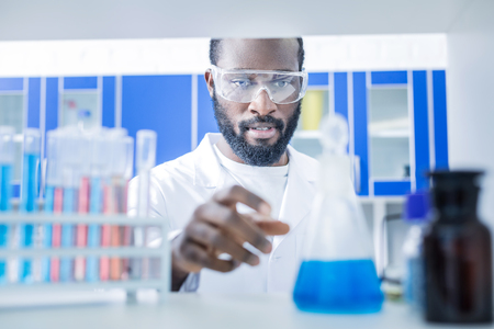 Scientific research. Serious nice smart man wearing protective glasses and looking at the flask while doing a scientific researchの写真素材