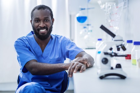Joyful mood. Happy pleasant male scientist sitting in the chair and looking at you while working in the laboratoryの写真素材