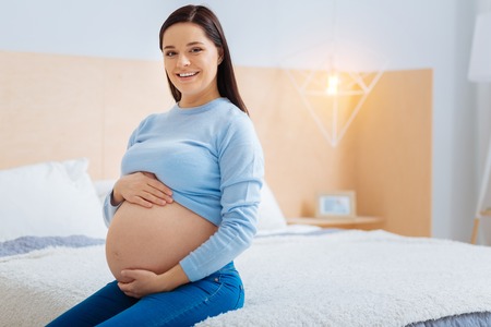 Home atmosphere. Young brunette woman touching her belly and keeping smile on her face while sitting in semi position on her bedの写真素材