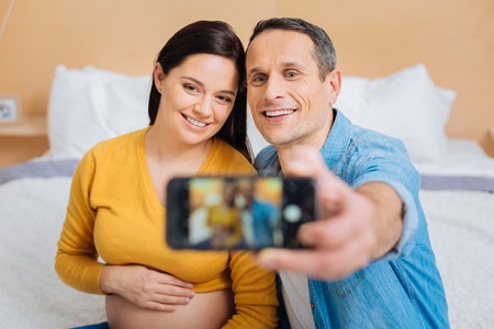 Pose on camera. Happy female person embracing her belly and sitting near her husband while taking family photoの写真素材