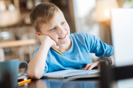 Smart child. Positive delighted smart boy holding his chin and smiling while looking at the laptop screenの写真素材