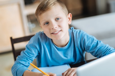 Studying at home. Positive cheerful smart boy smiling and looking at you while studying at home