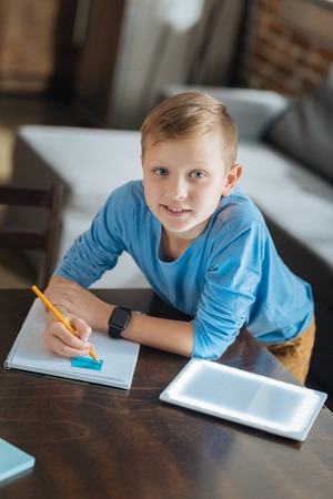 My home assignment. Nice pleasant intelligent boy standing near the table and writing in his copybook while doing his home assignmentの写真素材