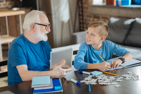 School lessons. Cheerful positive smart man holding a tablet and looking at his grandson while helping him with his school lessonsの写真素材