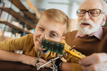 Magnifying glass. Nice pleasant curious boy leaning forward and looking into the magnifying glass while sitting at the table with his grandfatherの写真素材