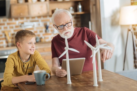Ecological energy. Smart nice aged man holding a tablet and pointing at the windmill models while teaching his grandson to care about environmentの写真素材