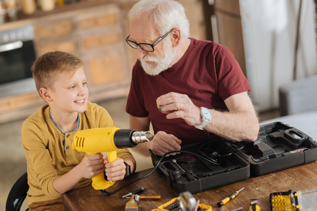 Male skills. Joyful nice positive boy sitting next to his grandfather and holding a drill while helping himの写真素材