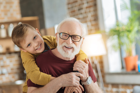 Warm relationships. Joyful nice loving boy standing behind his grandfather and hugging his while expressing his feelingsの写真素材