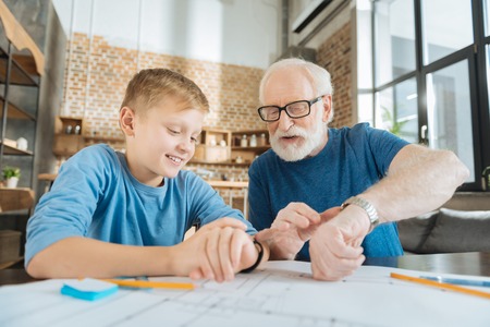 Technological differences. Positive nice cheerful grandfather and grandson sitting together and looking at their watches while comparing their technologyの写真素材