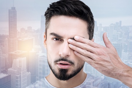 Be serious. Handsome male person keeping mouth opened and covering left eye while standing against urban backgroundの写真素材