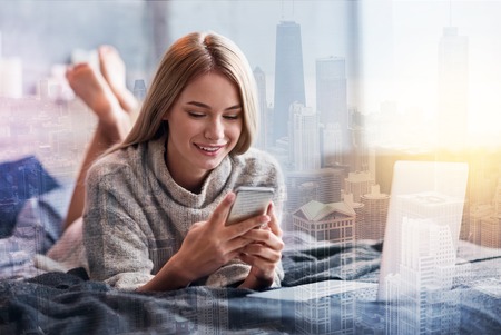 Social networks. Happy nice positive woman lying on her bed and holding her smartphone while using social networksの写真素材