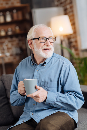Healthy drink. Pleasant senior man in eyeglasses sitting on the sofa and drinking tea from the big mug while looking into the distance and smilingの写真素材