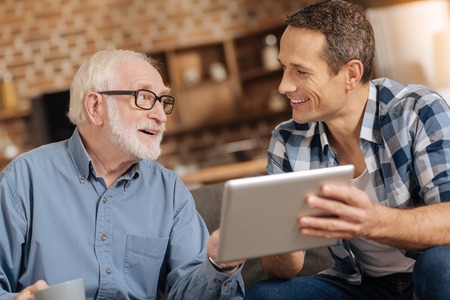 Exchanging opinions. Pleasant elderly man and his adult son sitting on the sofa together, reading a news article together and discussing itの写真素材