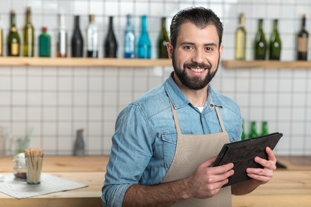 Convenient gadget. Positive emotional bearded waiter looking glad while standing with a modern...