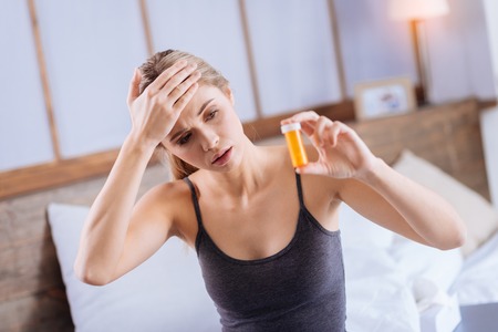 High blood heat. Petite young woman touching her forehead and looking at pills container in her hand while sitting on the bedの写真素材