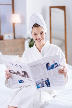 Recent news. Charming young woman sitting on the bed and reading a newspaper while wearing a towel turban and a bathrobe, having taken a showerの写真素材