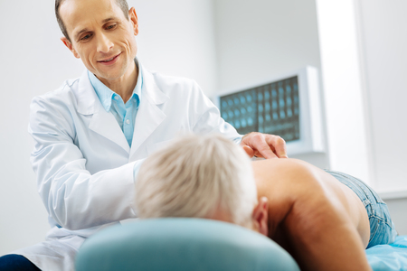 Physiotherapy center. Pleasant nice delighted doctor standing near his patient and smiling while doing a massage for himの写真素材