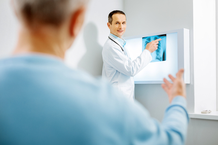 X ray diagnostics. Nice delighted male therapist pointing at the X ray photo and looking at his patient while putting a diagnosisの写真素材