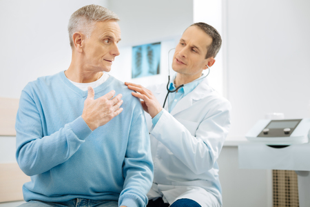 Professional osteopath. Smart handsome male doctor sitting behind his patient and wearing stethoscope while examining himの写真素材