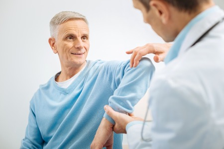 Problem with joints. Cheerful positive senior man bending his arm and letting the doctor check it while having a medical examinationの写真素材
