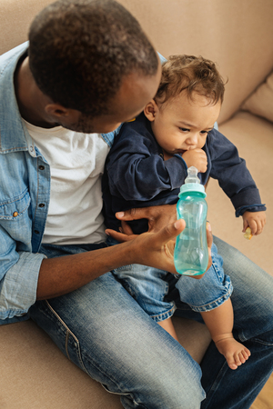 Feeding. Dark-haired caring afro-american father holding his little son and feeding him while sitting on the sofaの写真素材