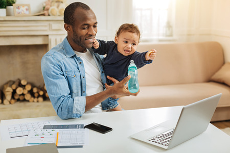 Tasty. Smiling loving young afro-american father holding his little son and feeding him while sitting at the table with his laptopの写真素材