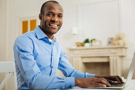 Working weekend. Handsome inspired afro-american man smiling and working on the laptop while sitting at the tableの写真素材
