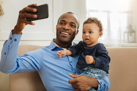 Look here. Handsome cheerful young afro-american daddy smiling and holding his little curly-haired son while taking selfiesの写真素材