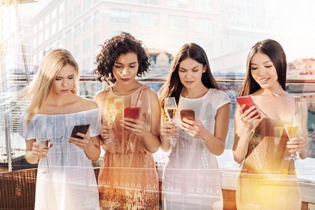 Social networking. Serious pleasant young women holding their smartphones and looking at their screens while standing togetherの写真素材