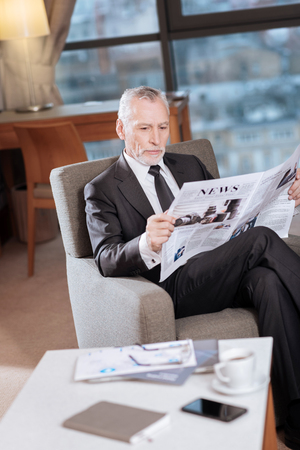 Last news. Experienced calm senior businessman  getting acquainted with news while relaxing in the armchair and looking downの写真素材