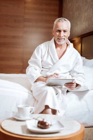 Excellent service. Senior enthusiastic cheerful man sitting on the bed carrying tablet and smilingの写真素材