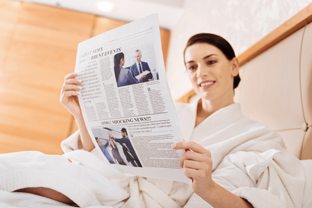 Daily news. Joyful optimistic gay woman scrutinizing newspaper  while holding it and wearing bathrobeの写真素材