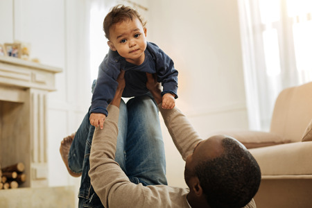 My love. Dark-haired bearded afro-american man playing with his cheerful young son while lying on the floor and holding the child in his armsの写真素材